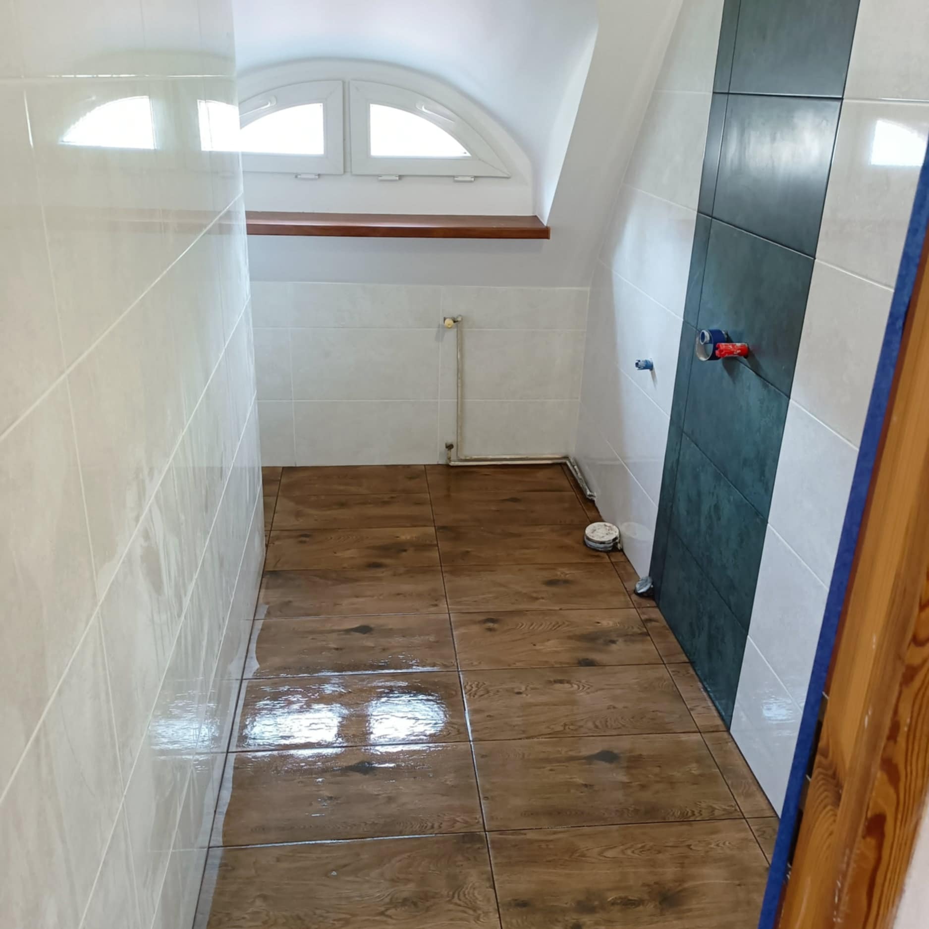 Nowoczesna łazienka po remoncie: brązowe płytki imitujące drewno na podłodze, jasne płytki na ścianach, ciemny pas płytek. Okno w kształcie łuku.