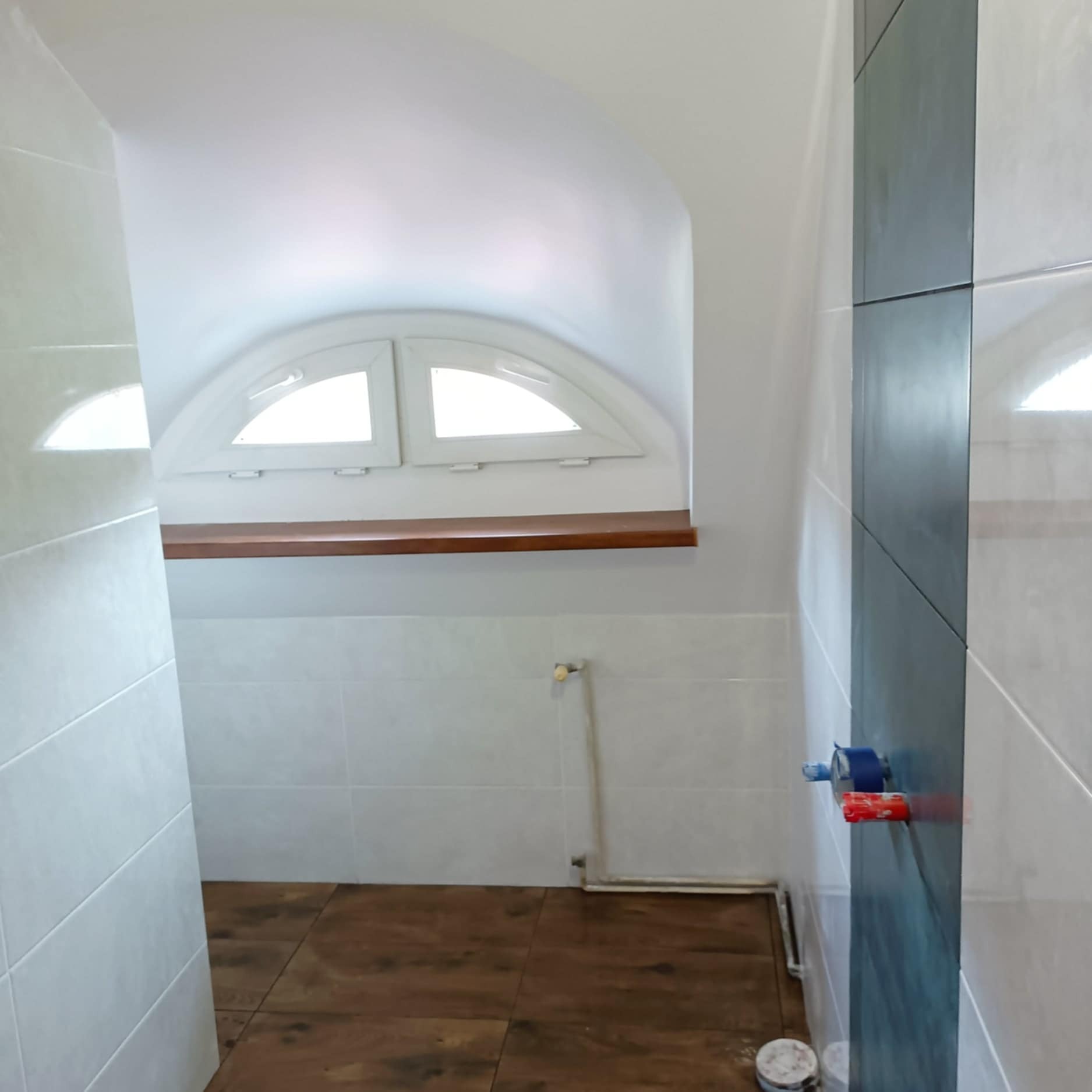 Wykończona łazienka z drewnopodobną podłogą i białymi płytkami. Nietypowe okno w kształcie półkola. Widoczne zawory na ścianie z ciemnymi płytkami.