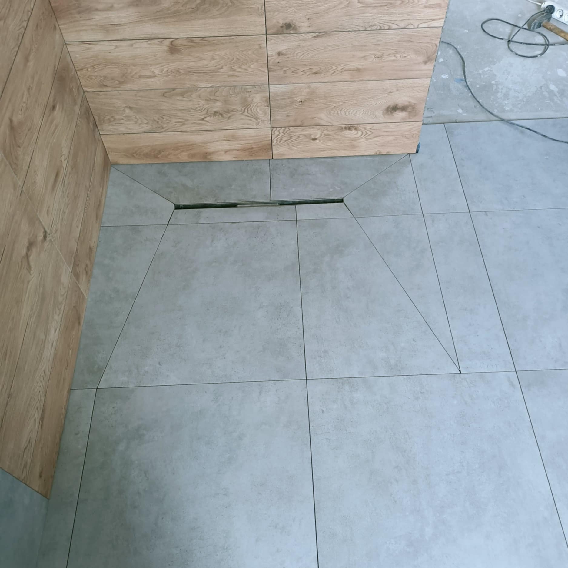 Wykończona podłoga w łazience z szarych płytek, z odpływem liniowym i drewnopodobną okładziną ściany. Widoczne narzędzia glazurnicze w tle.