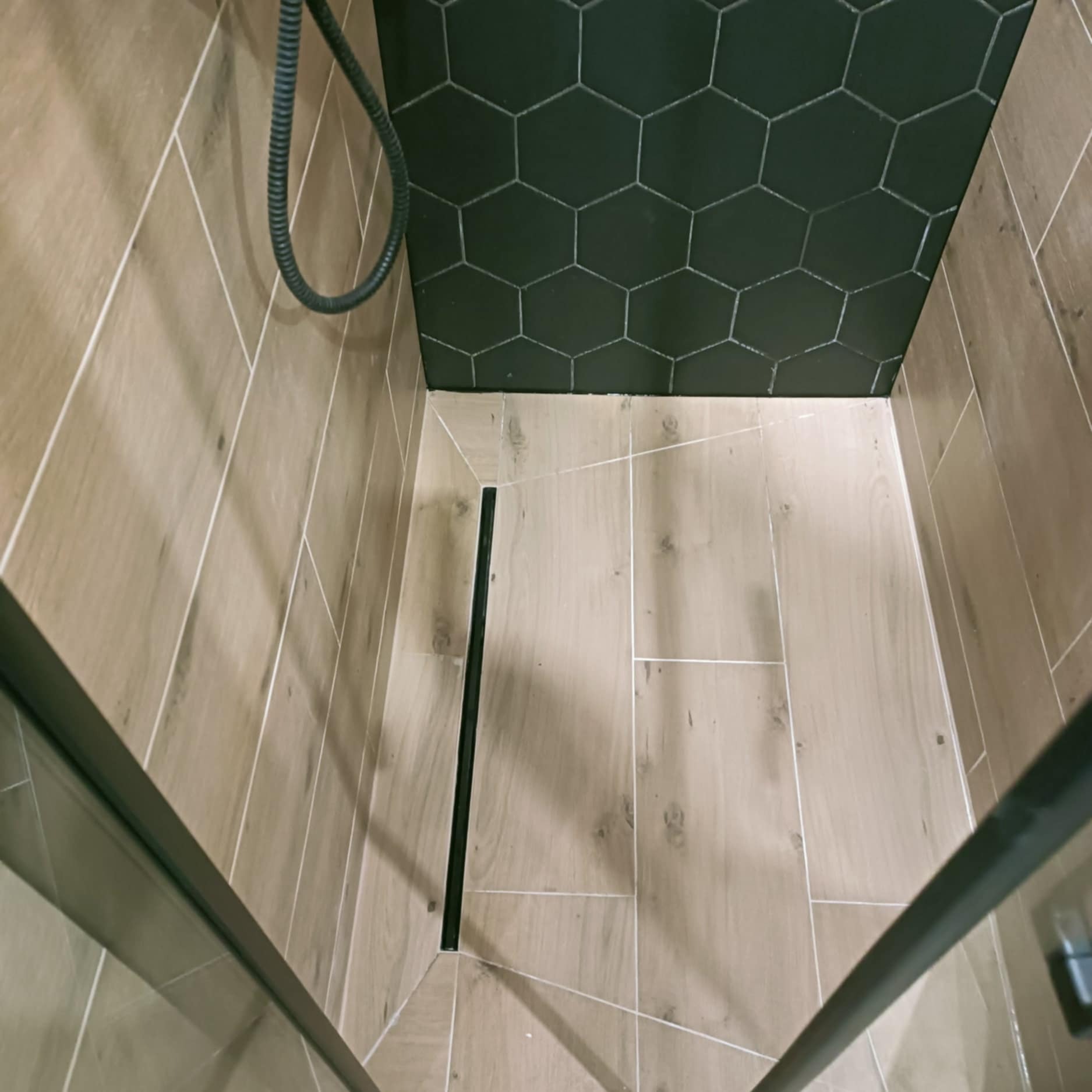 Wykończona kabina prysznicowa z odpływem liniowym, wyłożona płytkami imitującymi drewno. Na ścianie heksagonalne, grafitowe płytki.