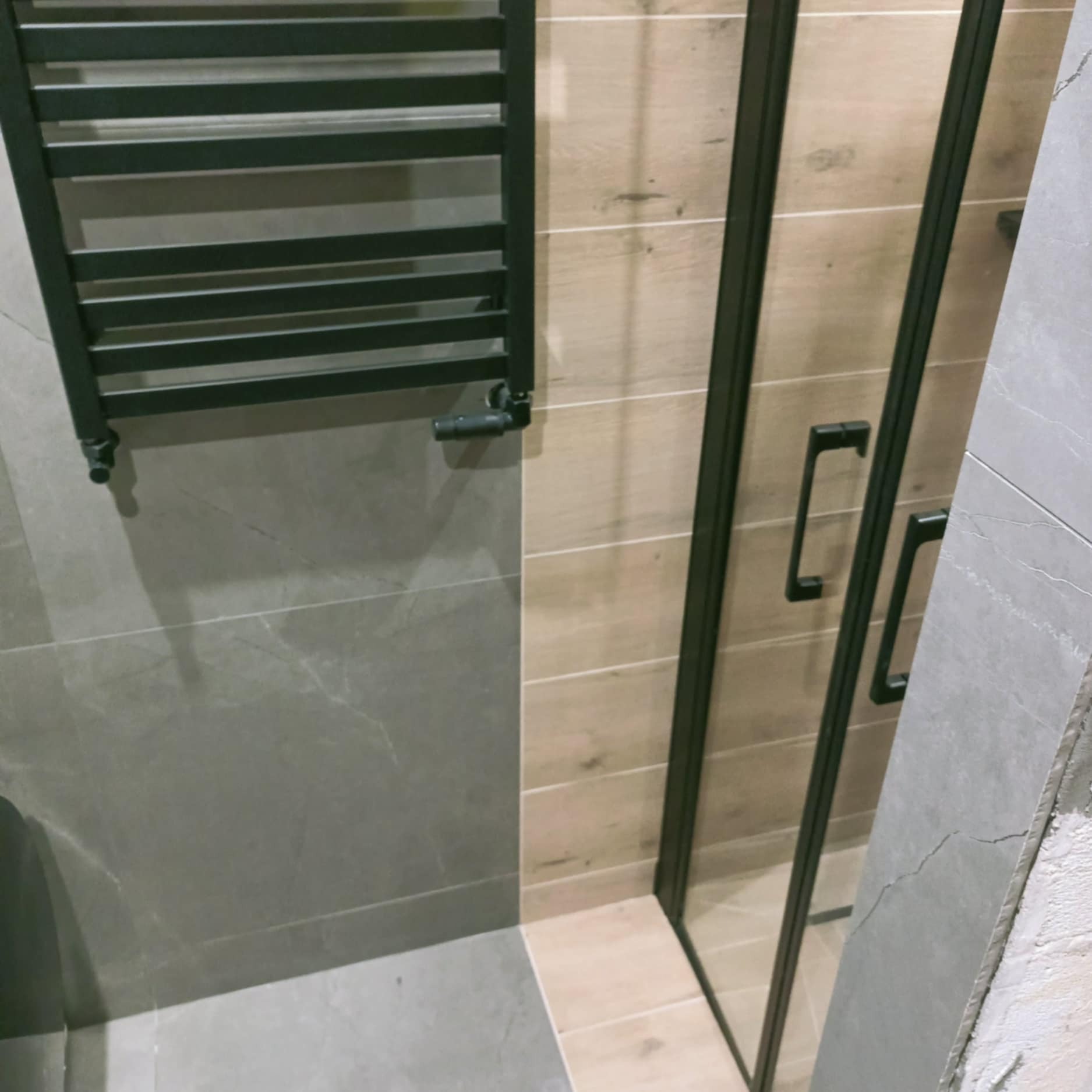 Nowoczesna łazienka po remoncie: grzejnik drabinkowy, kabina prysznicowa z czarnymi profilami, płytki imitujące drewno i szare płytki.