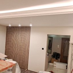 ARTBUD PRO Łukasz Sypnik - Wykończone wnętrze: białe ściany, dekoracyjne listwy sufitowe z oświetleniem LED, drewniana ścianka działowa z pionowych listew. Widoczne narzędzia i materiały budowlane.