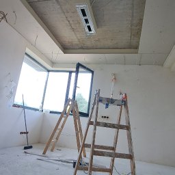 ARTBUD PRO Łukasz Sypnik - Wnętrze w trakcie prac: widoczne schody malarskie, okno, sufit z płyt kartonowo-gipsowych i instalacja oświetleniowa. W tle ściany przygotowane do dalszych prac wykończeniowych.