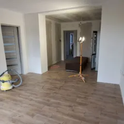 Wnętrze po szpachlowaniu i malowaniu ścian na biało, widoczny odkurzacz przemysłowy i lampa budowlana. Podłoga wyłożona panelami imitującymi drewno.