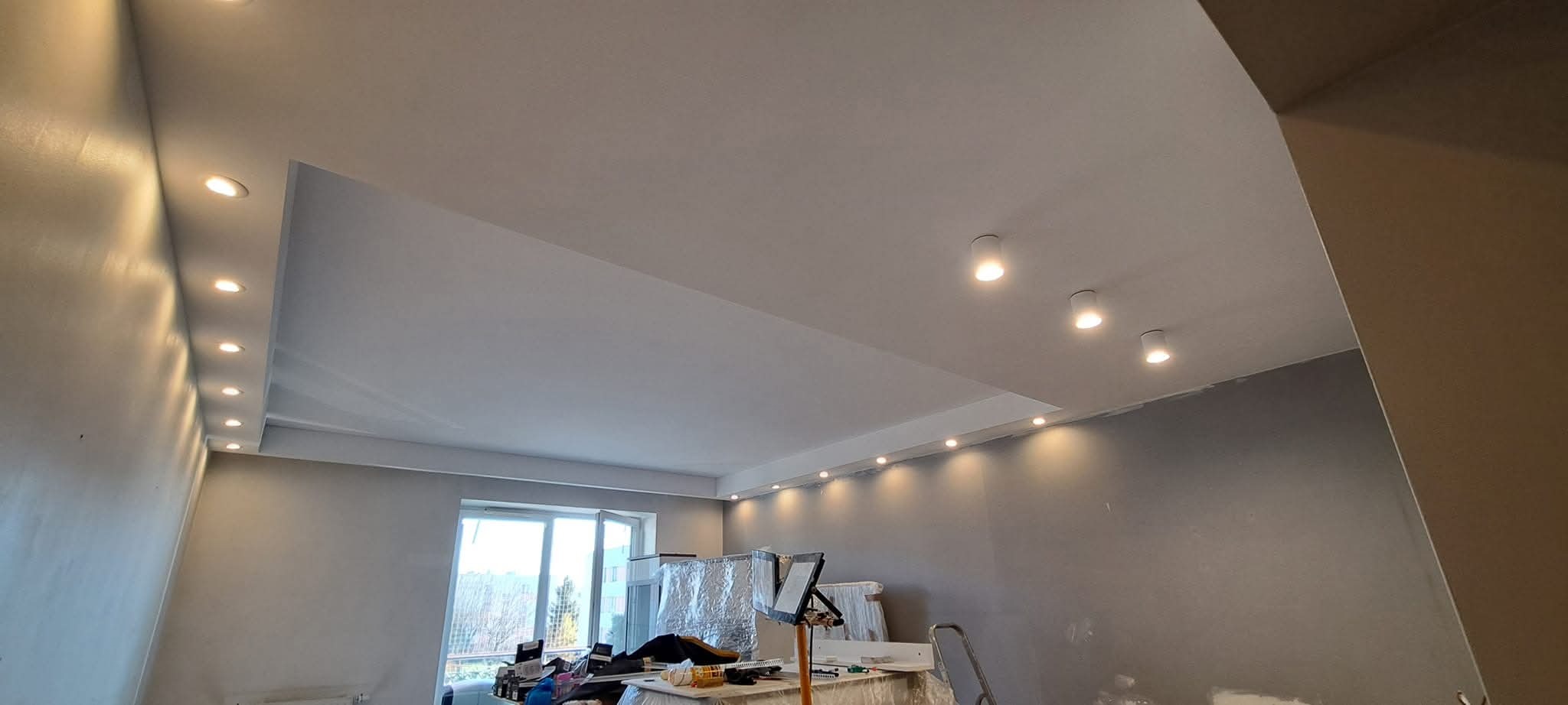 Wykończony sufit podwieszany z oświetleniem punktowym w szarym pokoju. Widoczne narzędzia i drabina malarska. Realizacja po remoncie.