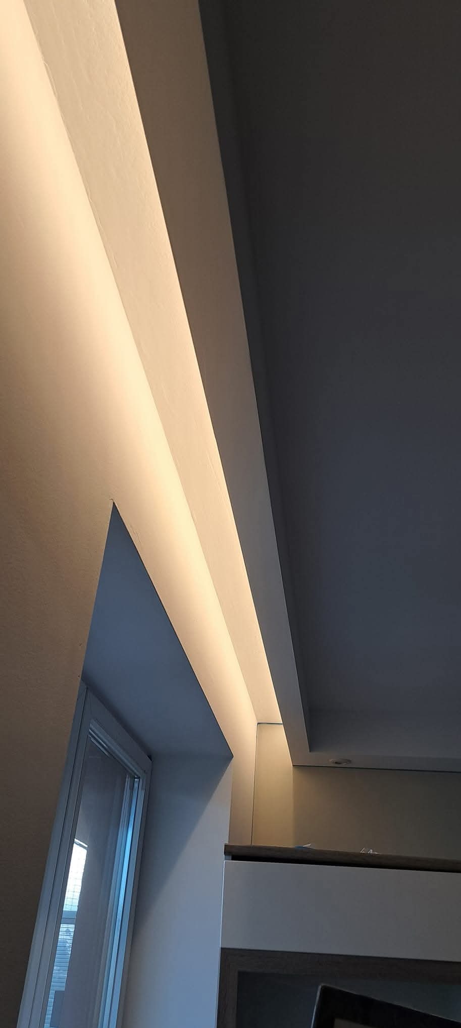 Nowoczesne oświetlenie sufitowe LED wnękowe, ciepłe światło podkreśla gładką powierzchnię ściany i sufitu. Widoczny fragment okna i mebli.