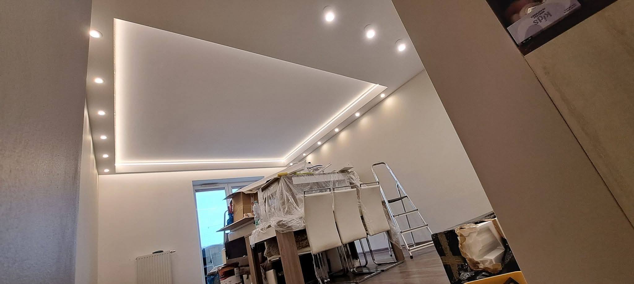 Wykończony sufit podwieszany z oświetleniem punktowym i LED w jasnym wnętrzu. Widoczne meble zabezpieczone folią i drabina.