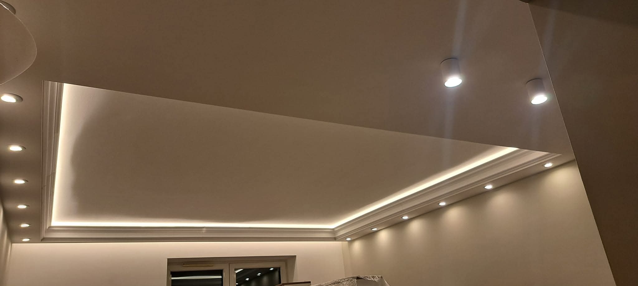 Nowoczesny sufit podwieszany z oświetleniem LED i dekoracyjną listwą gipsową. Widoczne punktowe lampy sufitowe i fragment okna.