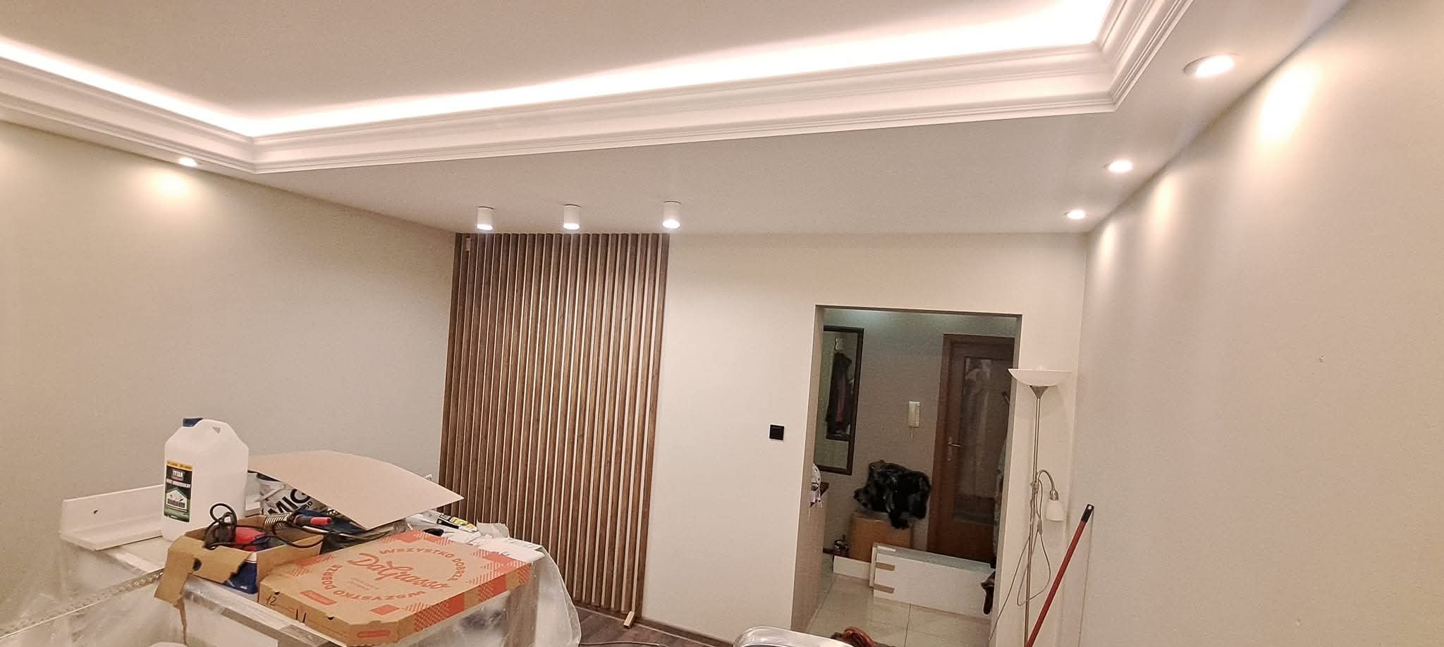 Wykończone wnętrze: białe ściany, dekoracyjne listwy sufitowe z oświetleniem LED, drewniana ścianka działowa z pionowych listew. Widoczne narzędzia i materiały budowlane.