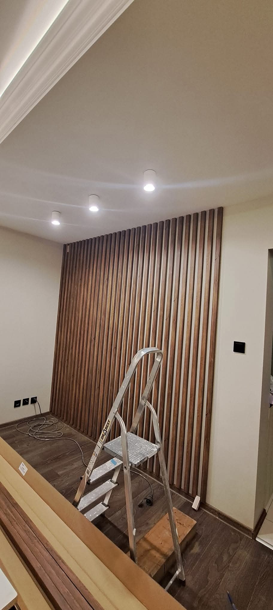 Montaż drewnianej boazerii pionowej na ścianie. Widoczna drabina i listwy podłogowe. Nowoczesny design wnętrza z oświetleniem punktowym.