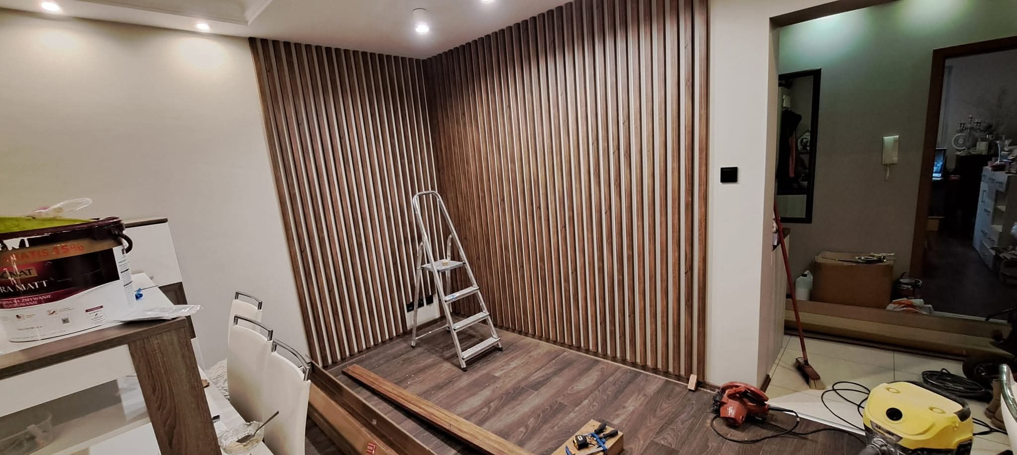 Drewniane lamele na ścianie w trakcie montażu, drabina, narzędzia i farba widoczne. Podłoga drewniana, oświetlenie sufitowe, wnętrze w trakcie prac wykończeniowych.