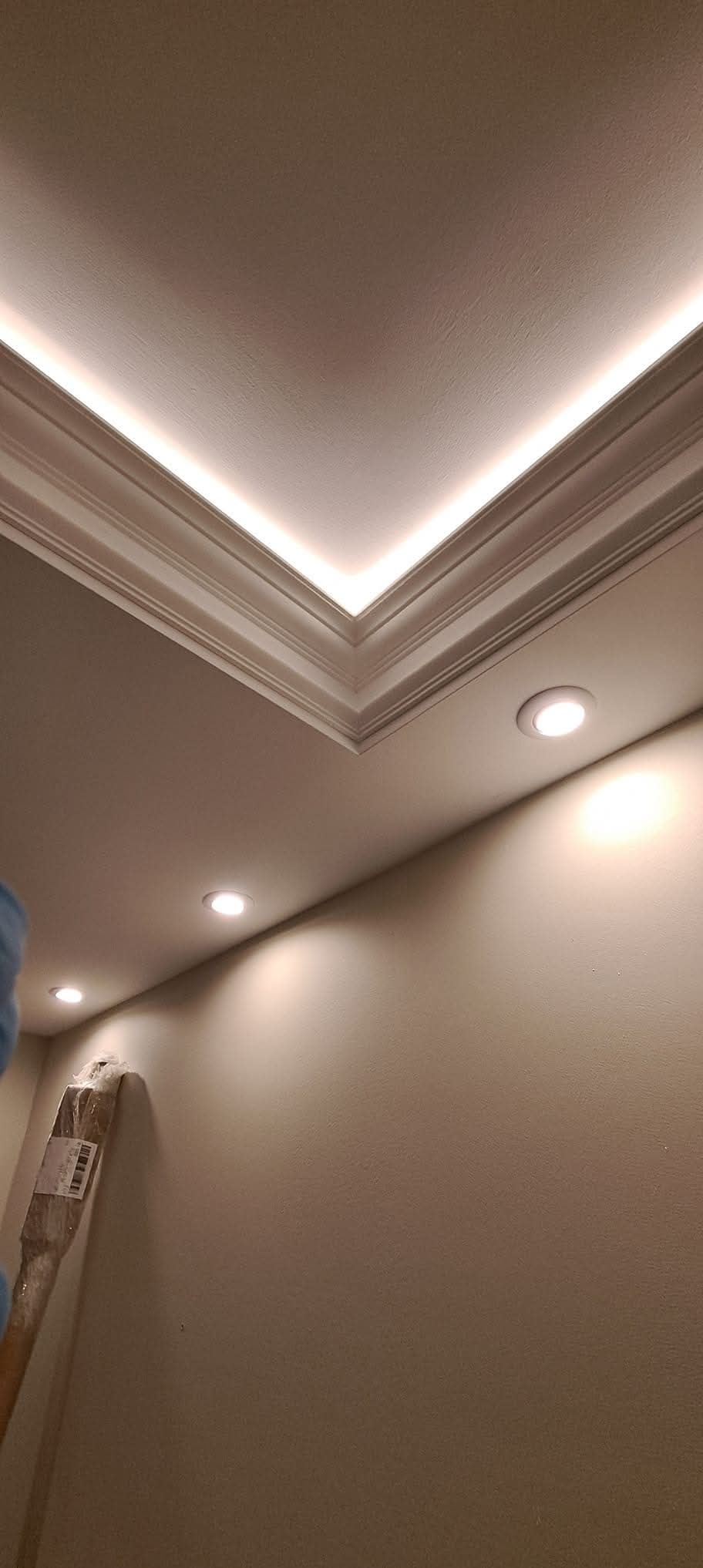 Wykończony sufit z oświetleniem LED w listwach i punktowymi lampami. Ściana pomalowana na beżowo, widoczny fragment drabiny malarskiej.