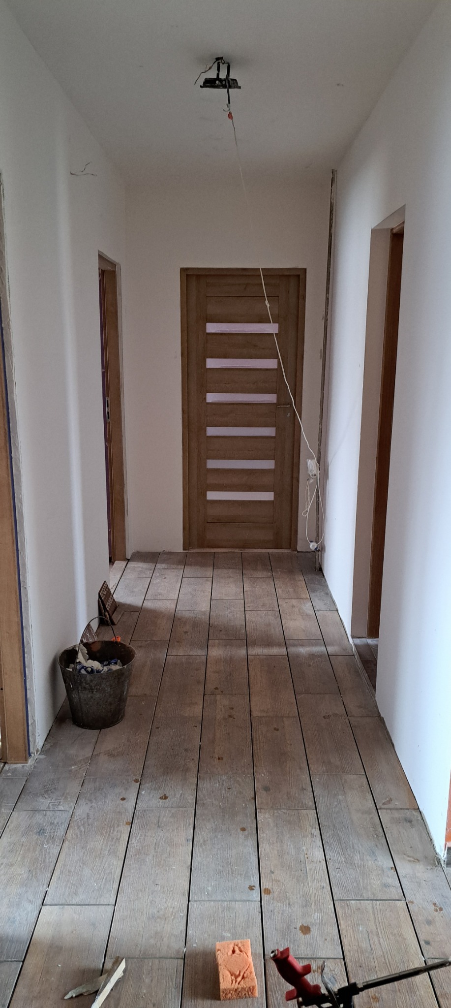 Korytarz w trakcie wykańczania: świeżo pomalowane ściany, drewniana podłoga z narzędziami, nowe drzwi z poziomymi przeszkleniami.