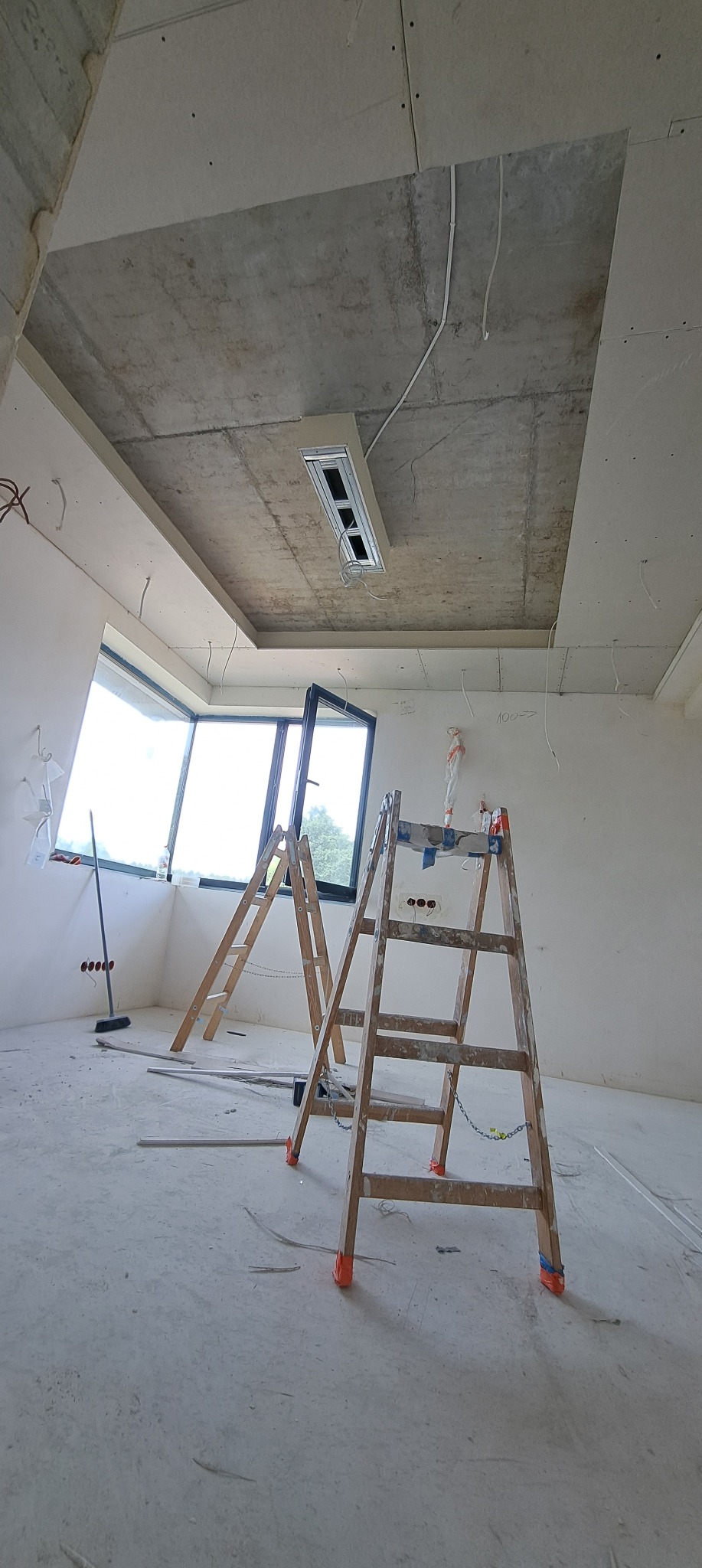 Wnętrze w trakcie prac: widoczne schody malarskie, okno, sufit z płyt kartonowo-gipsowych i instalacja oświetleniowa. W tle ściany przygotowane do dalszych prac wykończeniowych.