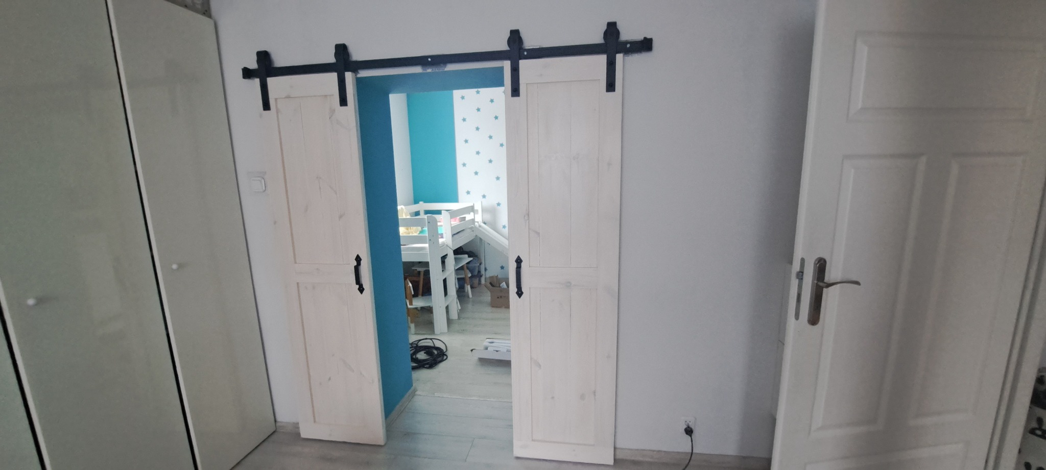 Białe, drewniane drzwi przesuwne w stylu rustykalnym na czarnym szynoprzewodzie, widok na pokój dziecięcy z łóżkiem i turkusową ścianą. Dodatkowe drzwi po prawej.