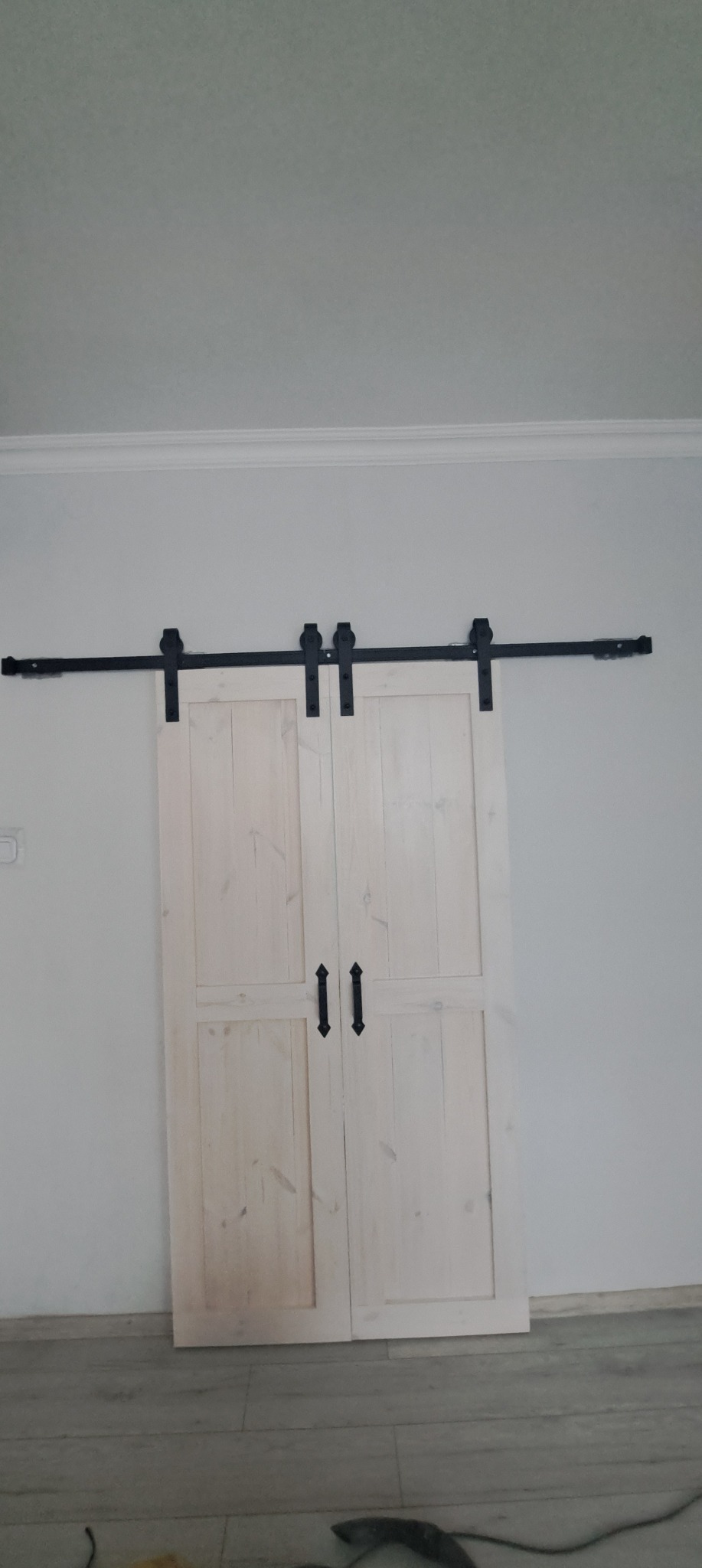 Jasne, drewniane drzwi przesuwne w stylu rustykalnym z czarnymi okuciami, zamontowane na szynie w jasnym wnętrzu. Widoczne detale wykończenia i faktura drewna.