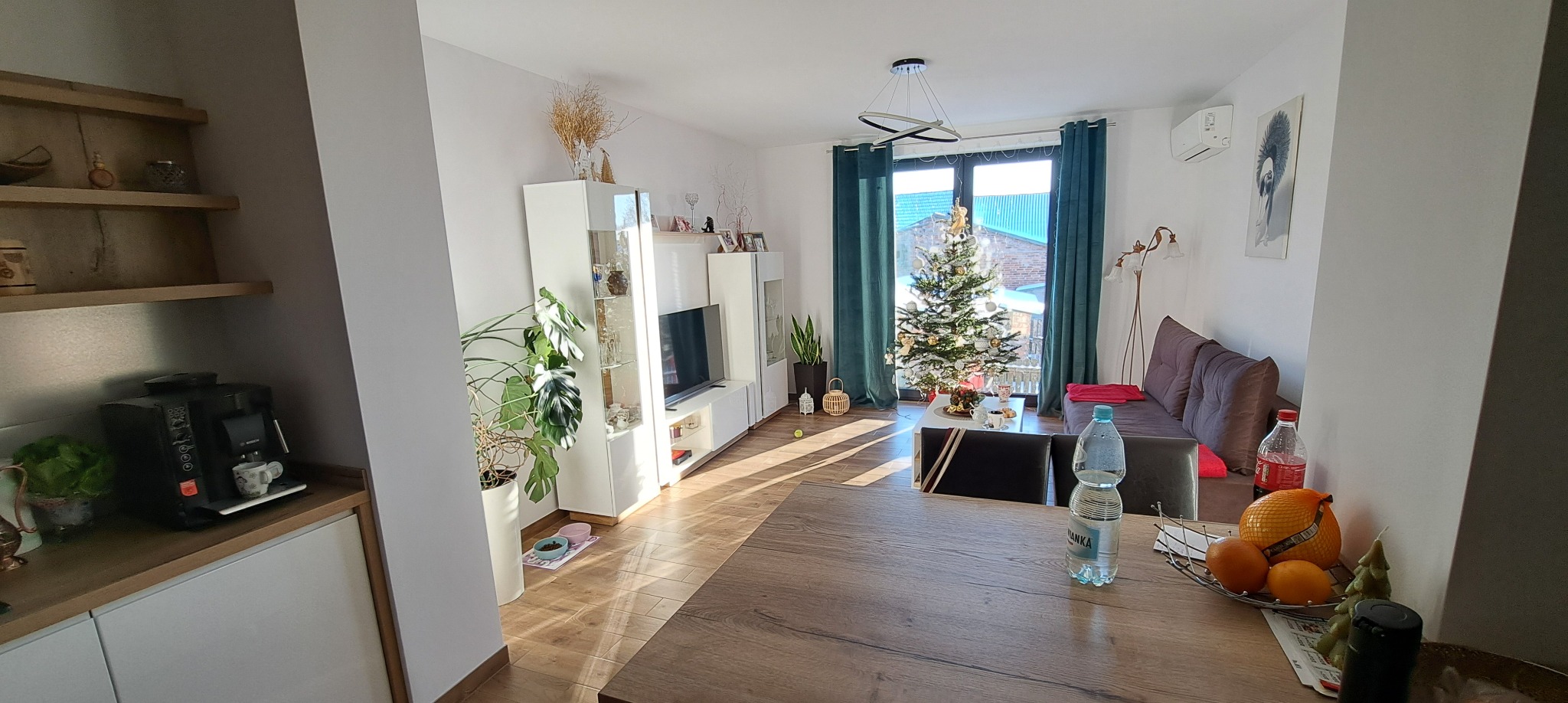 Jasne, przestronne wnętrze salonu z choinką i elementami dekoracyjnymi, widok z perspektywy kuchni, drewniana podłoga i meble w odcieniach bieli i brązu, nowoczesne oświetlenie.