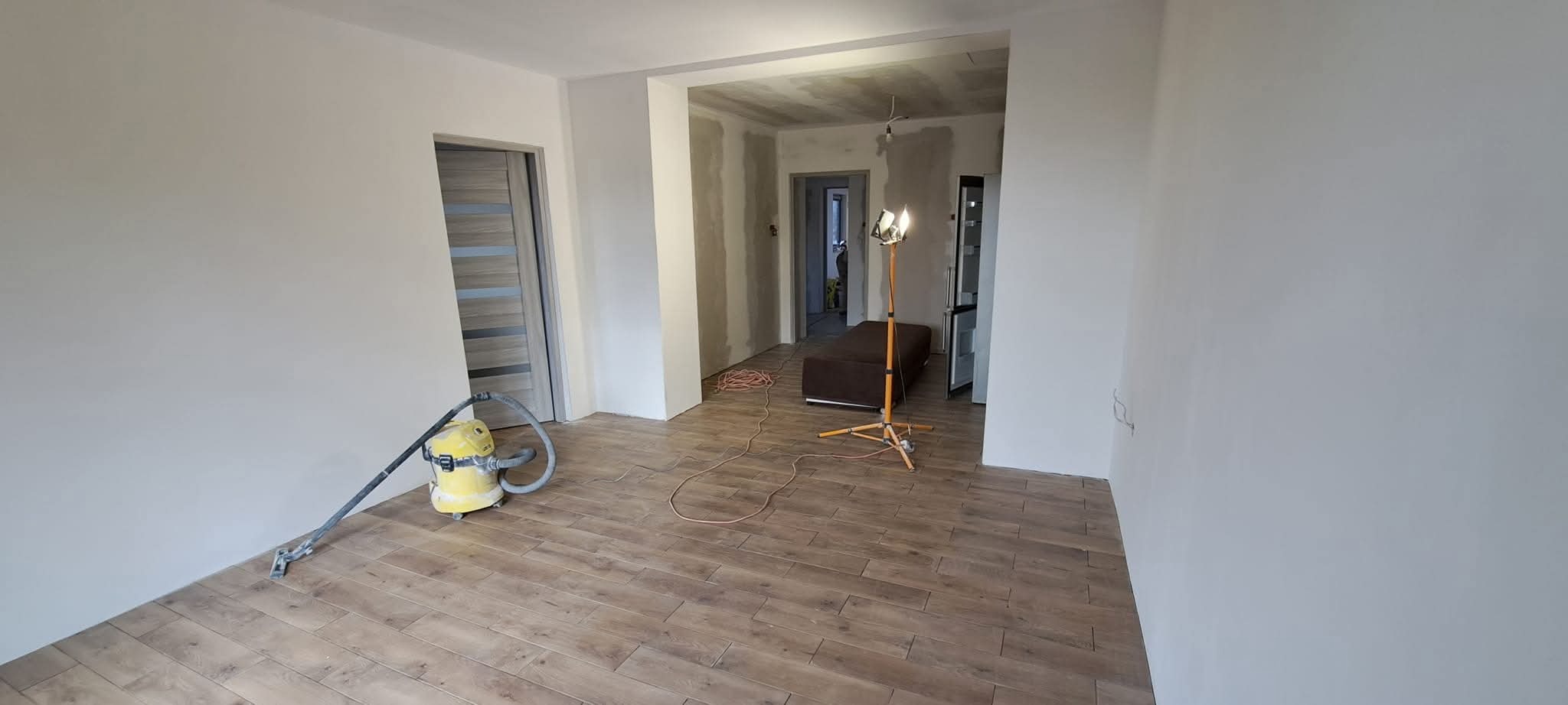 Wnętrze po szpachlowaniu i malowaniu ścian na biało, widoczny odkurzacz przemysłowy i lampa budowlana. Podłoga wyłożona panelami imitującymi drewno.