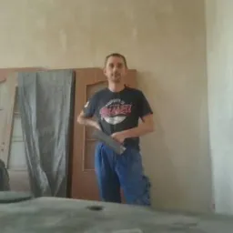 Mężczyzna z szpachelką na tle ściany przed malowaniem, obok drzwi. Przygotowania do szpachlowania ścian w pomieszczeniu, widoczne narzędzia pracy.