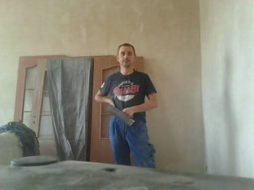 Mężczyzna z szpachelką na tle ściany przed malowaniem, obok drzwi. Przygotowania do szpachlowania ścian w pomieszczeniu, widoczne narzędzia pracy.