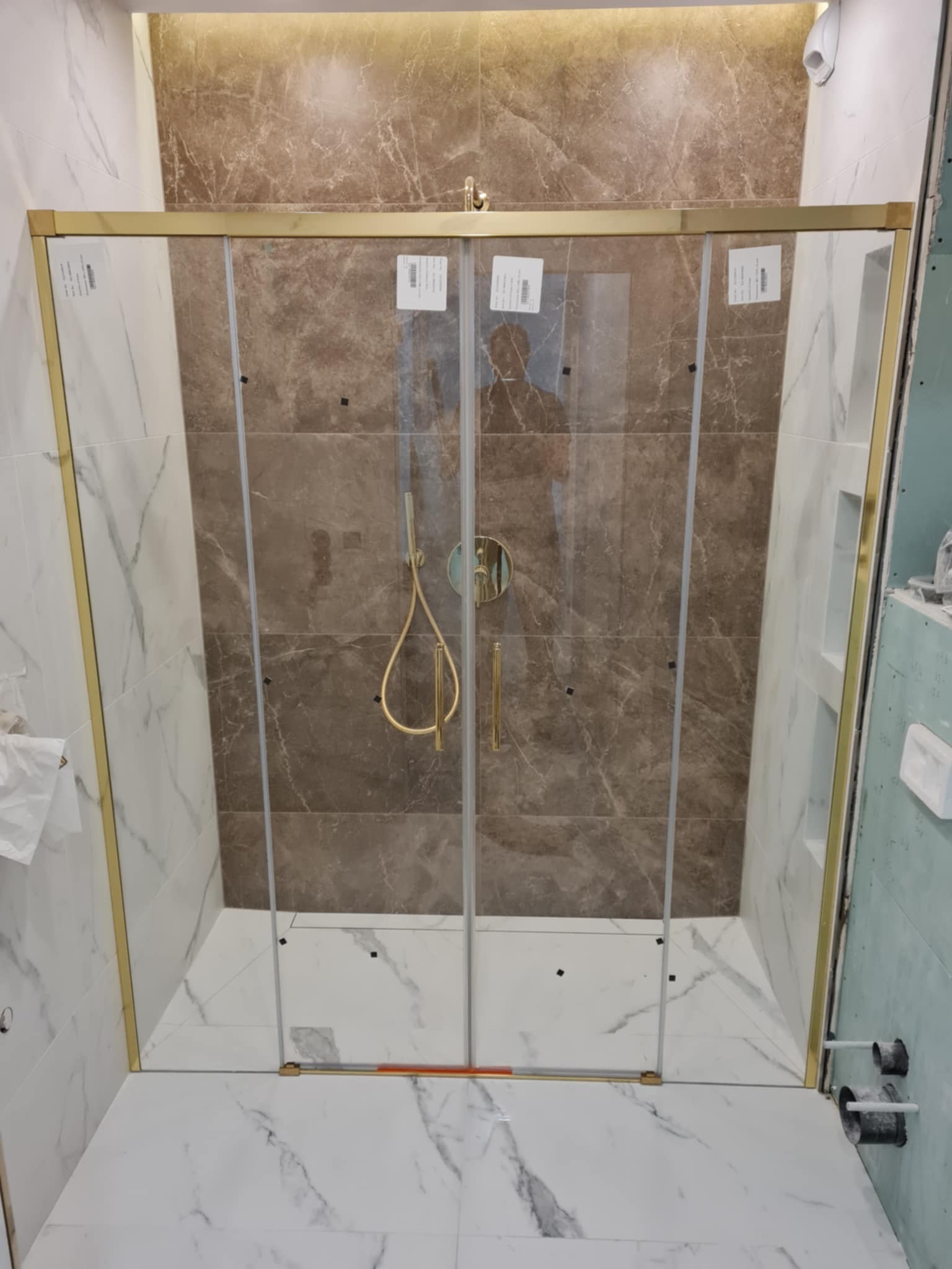 Kabina prysznicowa z złotymi profilami na tle marmurowych płytek w odcieniach bieli i brązu. Widoczne fragmenty instalacji wodnej i niedokończone wykończenie ściany.
