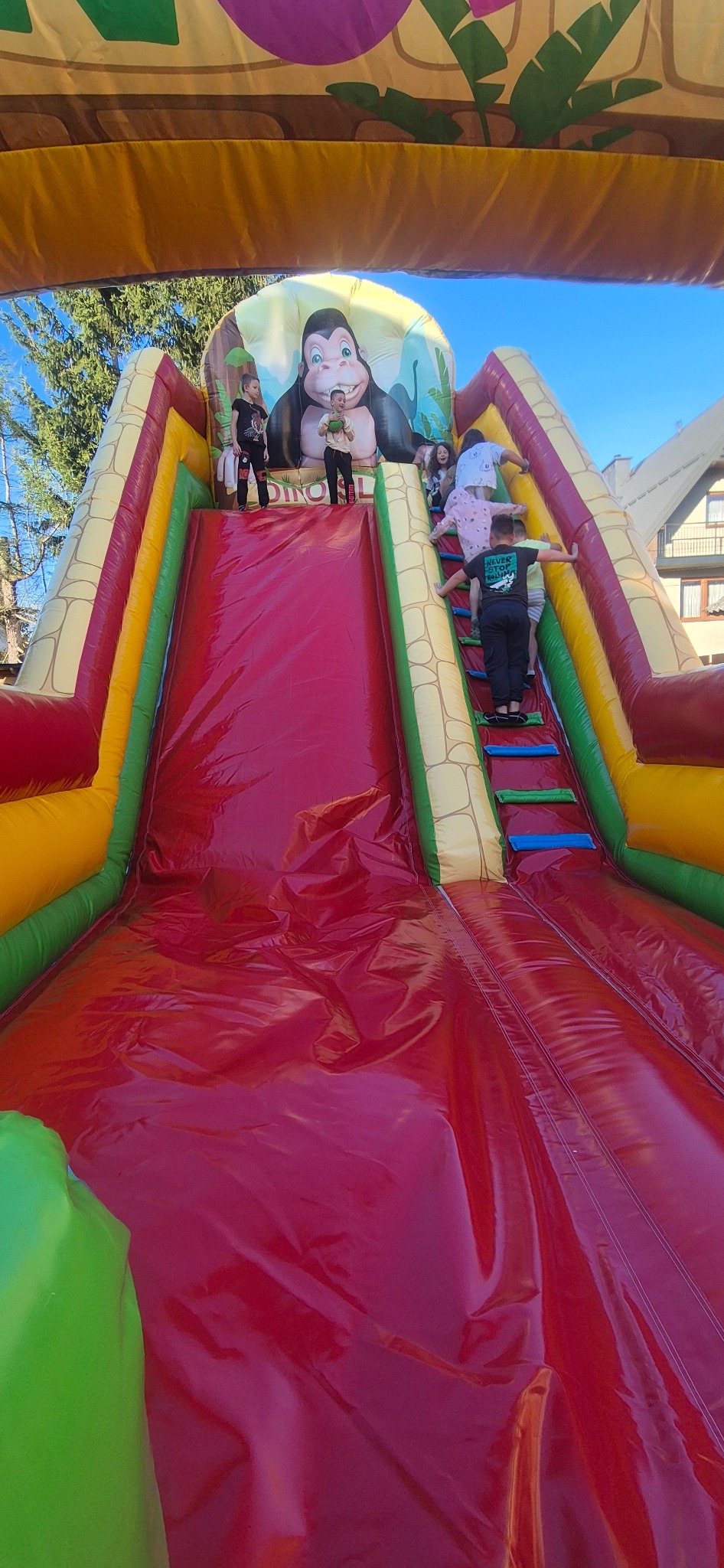 Kolorowa dmuchana zjeżdżalnia z motywem goryla i wspinaczką, na której bawi się grupa dzieci w słoneczny dzień. Atrakcja plenerowa.