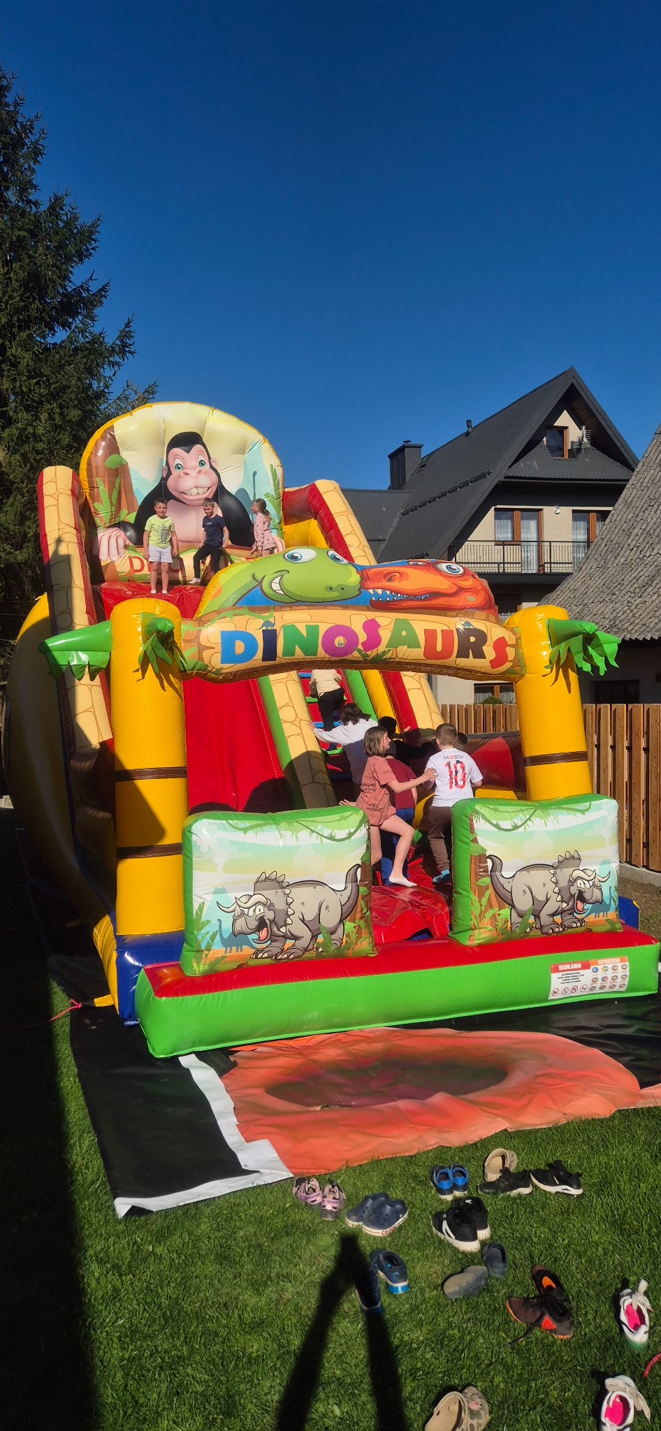 Kolorowy, duży dmuchany zamek z motywem dinozaurów i goryla, na którym bawi się grupa dzieci w słoneczny dzień. Na trawie leżą buty.