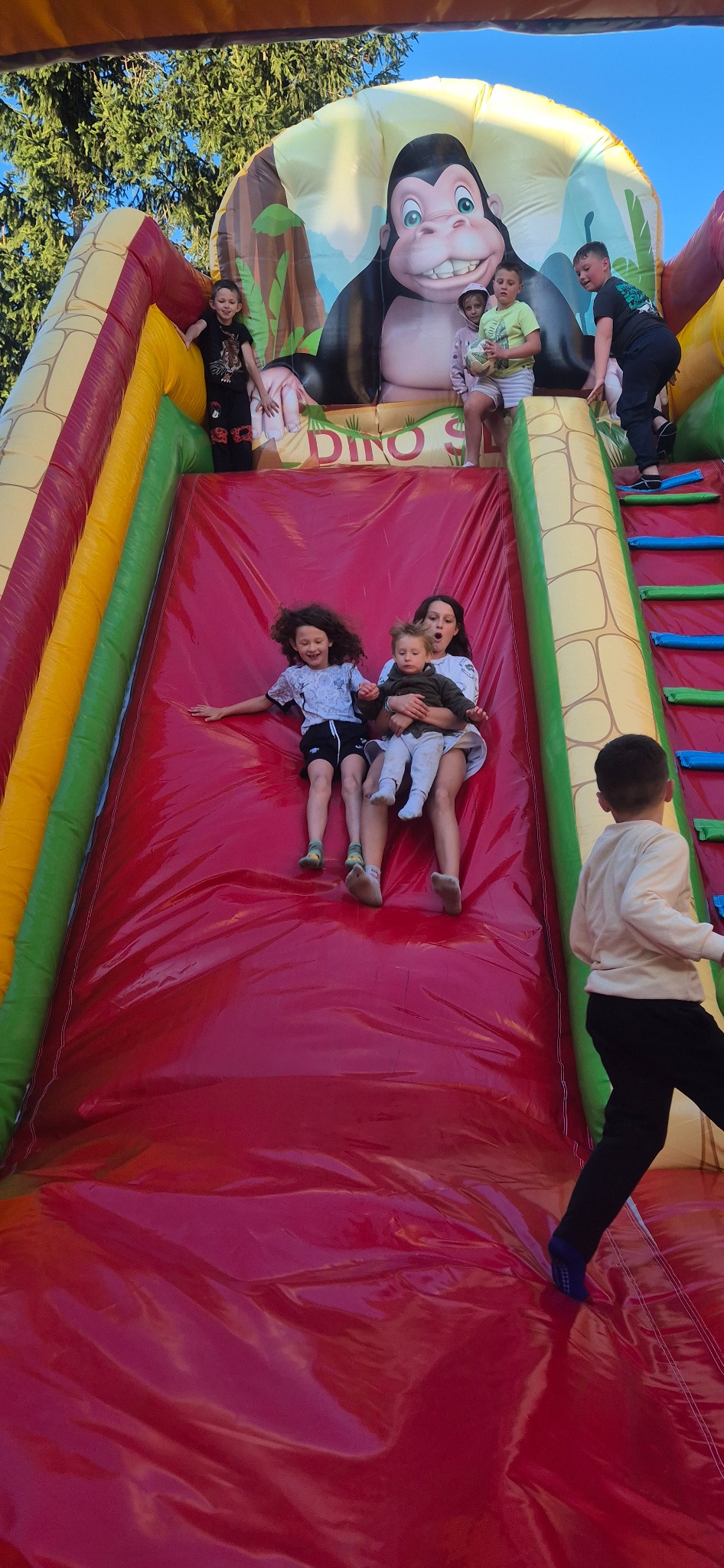 Dzieci zjeżdżają z dmuchanej zjeżdżalni z motywem goryla. Zjeżdżalnia ma czerwony ślizg i kolorowe boki. Widoczne drabinki i inne dzieci na górze.