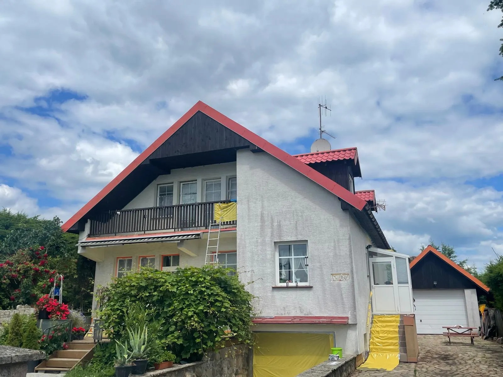 Dom z białymi ścianami i czerwonym dachem w trakcie przygotowań do malowania elewacji, zabezpieczony folią i z drabiną opartą o balkon.