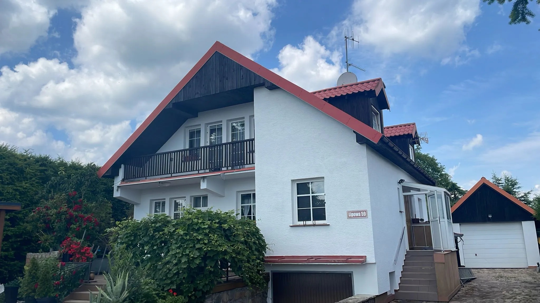 Biały dom z czerwonym dachem i ciemnym drewnianym wykończeniem, otoczony zielenią. Widoczny balkon z czarną balustradą. Tabliczka z adresem 'Lipowa 59'.