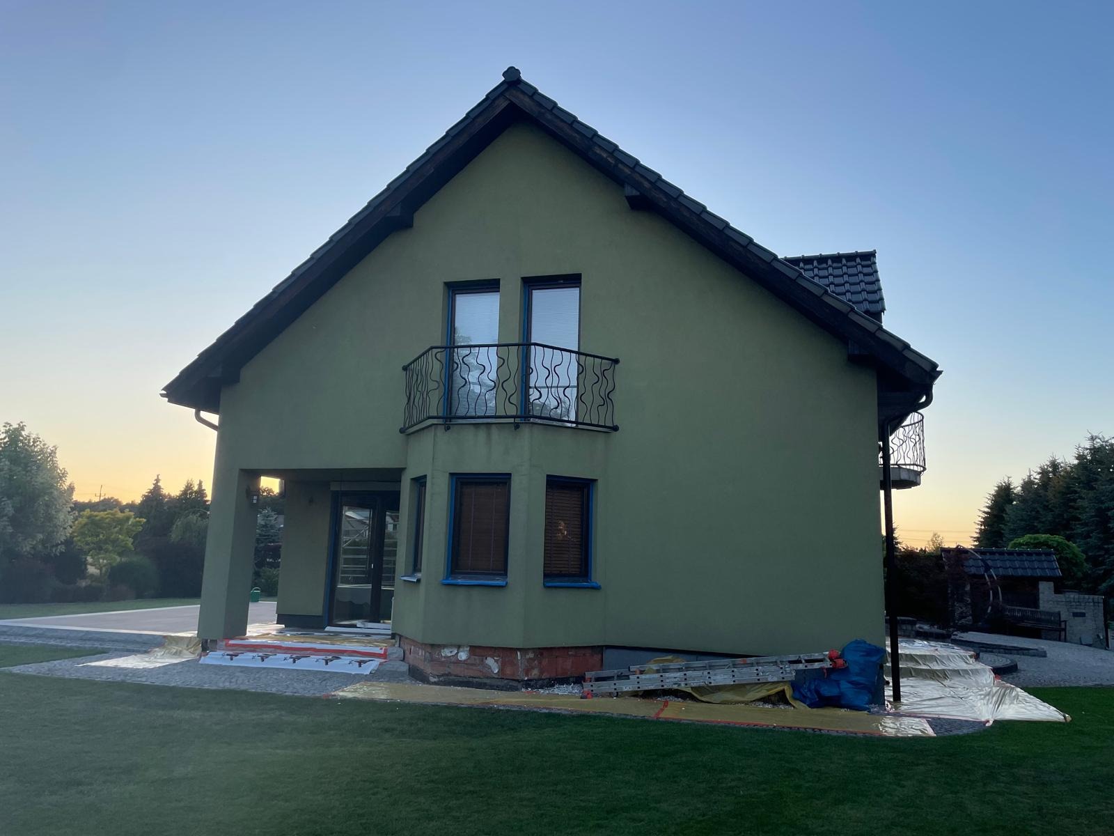 Dom z zieloną elewacją, widoczne przygotowania do prac: folie ochronne i drabina. Balustrady balkonowe, okna z roletami. Ujęcie z niskiego kąta, wieczorne światło.