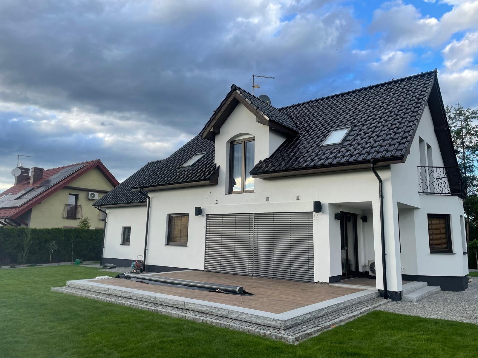 Biały dom jednorodzinny z czarnym dachem z dachówki, drewnianym tarasem i szarymi roletami zewnętrznymi, widok z zadbanego trawnika w pochmurny dzień.