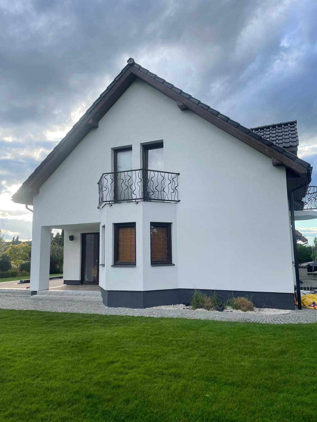 Biała elewacja domu jednorodzinnego z balkonem i ciemną podmurówką, widok z poziomu trawnika w pochmurny dzień. W tle widoczne drzewa i szare niebo.
