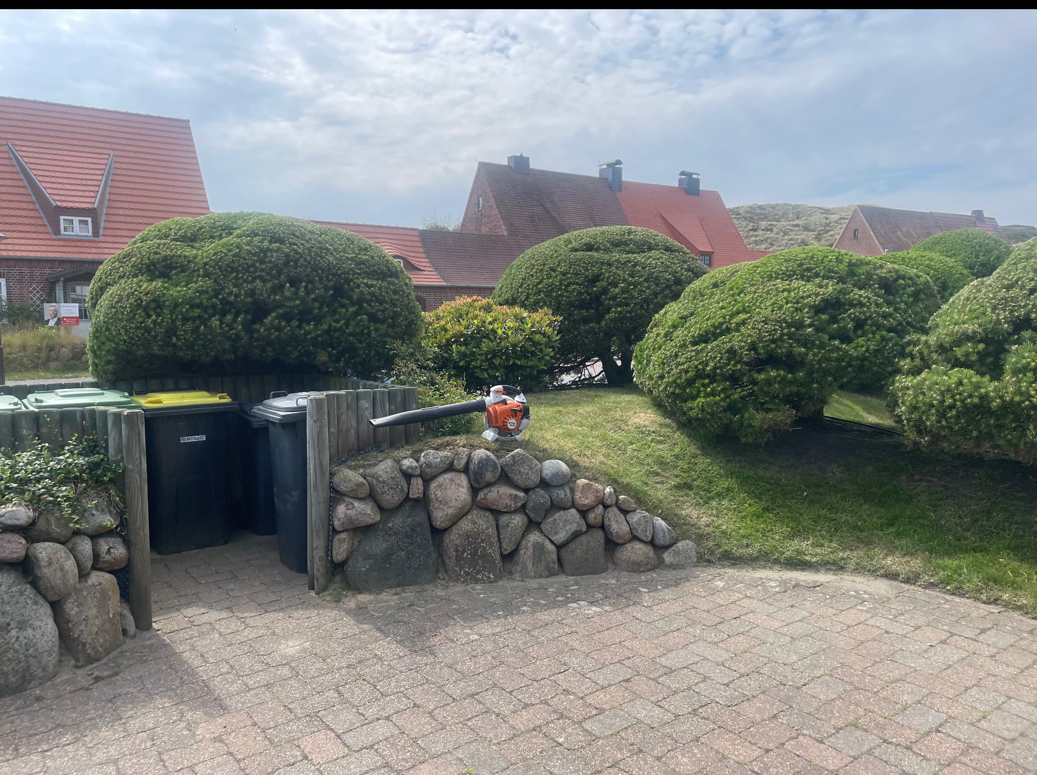Kształtne, kuliste krzewy w ogrodzie z kostki brukowej, z dmuchawą do liści Stihl na kamiennym murku. W tle domy z czerwonymi dachami i zielone pojemniki na śmieci.