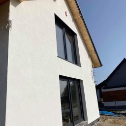 Tomik-BUD - Ściana domu z nową, jasną elewacją i czarnymi oknami, widok z dołu. Widoczne drewniane elementy dachu i fragment sąsiedniego budynku.