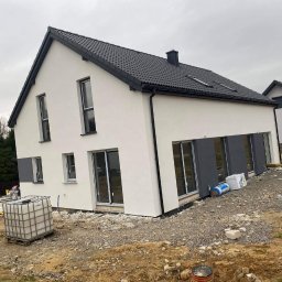 Tomik-BUD - Biały dom z szarą stolarką okienną i grafitowym dachem. Widoczne materiały budowlane na posesji. Dom po tynkowaniu, przed zagospodarowaniem terenu.