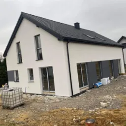 Biały dom z szarą stolarką okienną i grafitowym dachem. Widoczne materiały budowlane na posesji. Dom po tynkowaniu, przed zagospodarowaniem terenu.