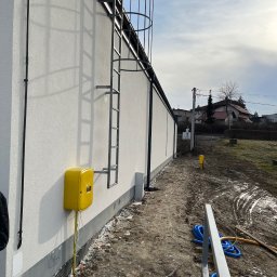 Tomik-BUD - Biała elewacja budynku z żółtą skrzynką gazową i drabinką w Żywcu. Widoczne elementy instalacji i teren wokół budynku. Teksturowana powierzchnia elewacji.