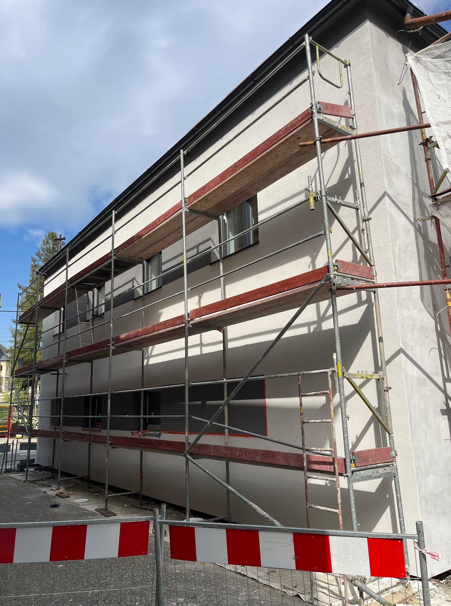 Elewacja budynku w trakcie prac wykończeniowych, widoczne rusztowania i świeżo nałożony tynk strukturalny. Budynek dwukondygnacyjny z oknami.