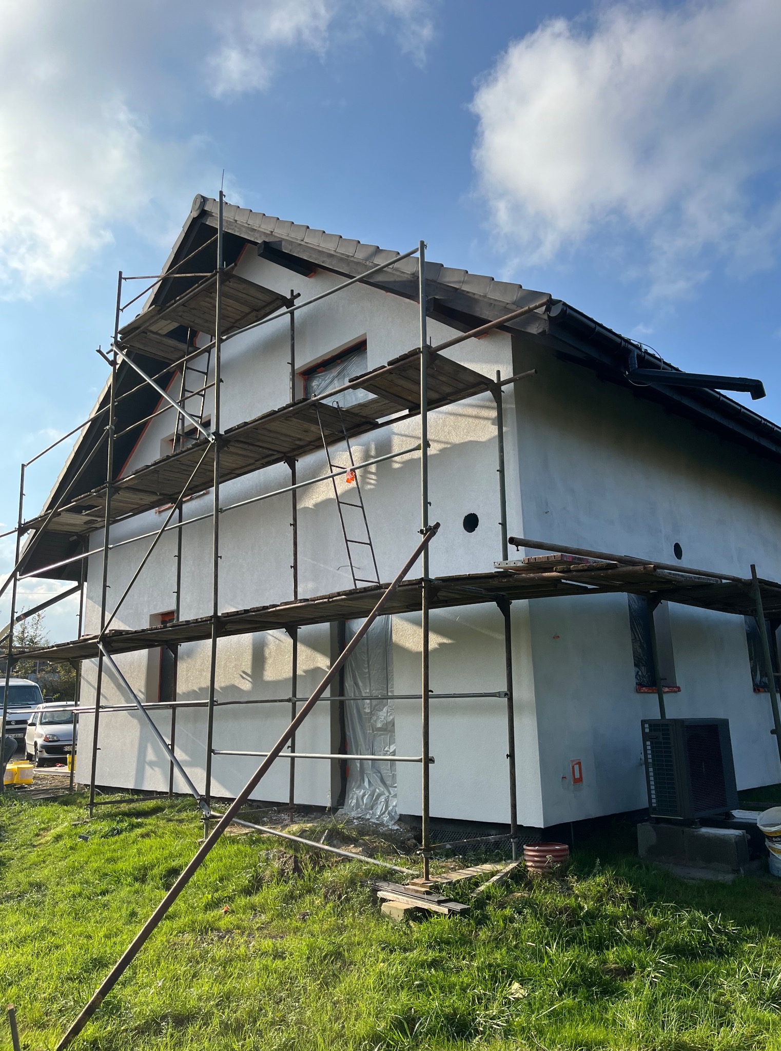 Dom w trakcie ocieplania, widoczne rusztowania i biała elewacja. Okna zabezpieczone folią, na trawie wiadro z farbą. Słoneczny dzień.