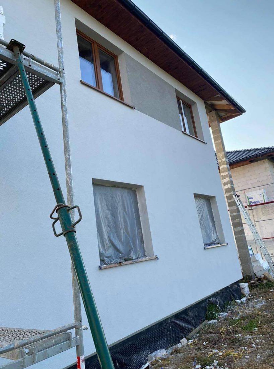 Dom po nałożeniu białego tynku, z widocznymi rusztowaniami i oknami zabezpieczonymi folią, przygotowany do dalszych prac wykończeniowych.