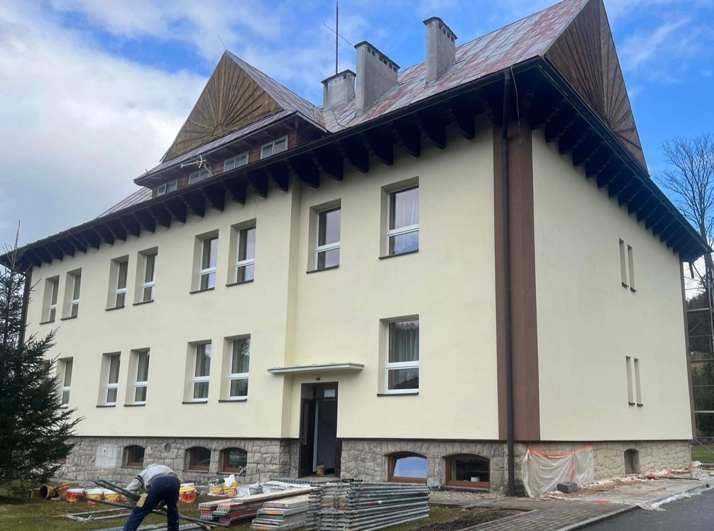 Odnowiony budynek z kremową elewacją i drewnianym wykończeniem dachu. Na pierwszym planie widoczne materiały budowlane i fragment pracownika.