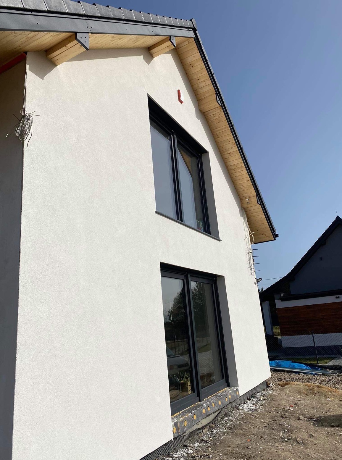 Ściana domu z nową, jasną elewacją i czarnymi oknami, widok z dołu. Widoczne drewniane elementy dachu i fragment sąsiedniego budynku.