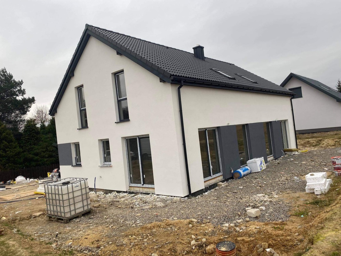 Biały dom z szarą stolarką okienną i grafitowym dachem. Widoczne materiały budowlane na posesji. Dom po tynkowaniu, przed zagospodarowaniem terenu.