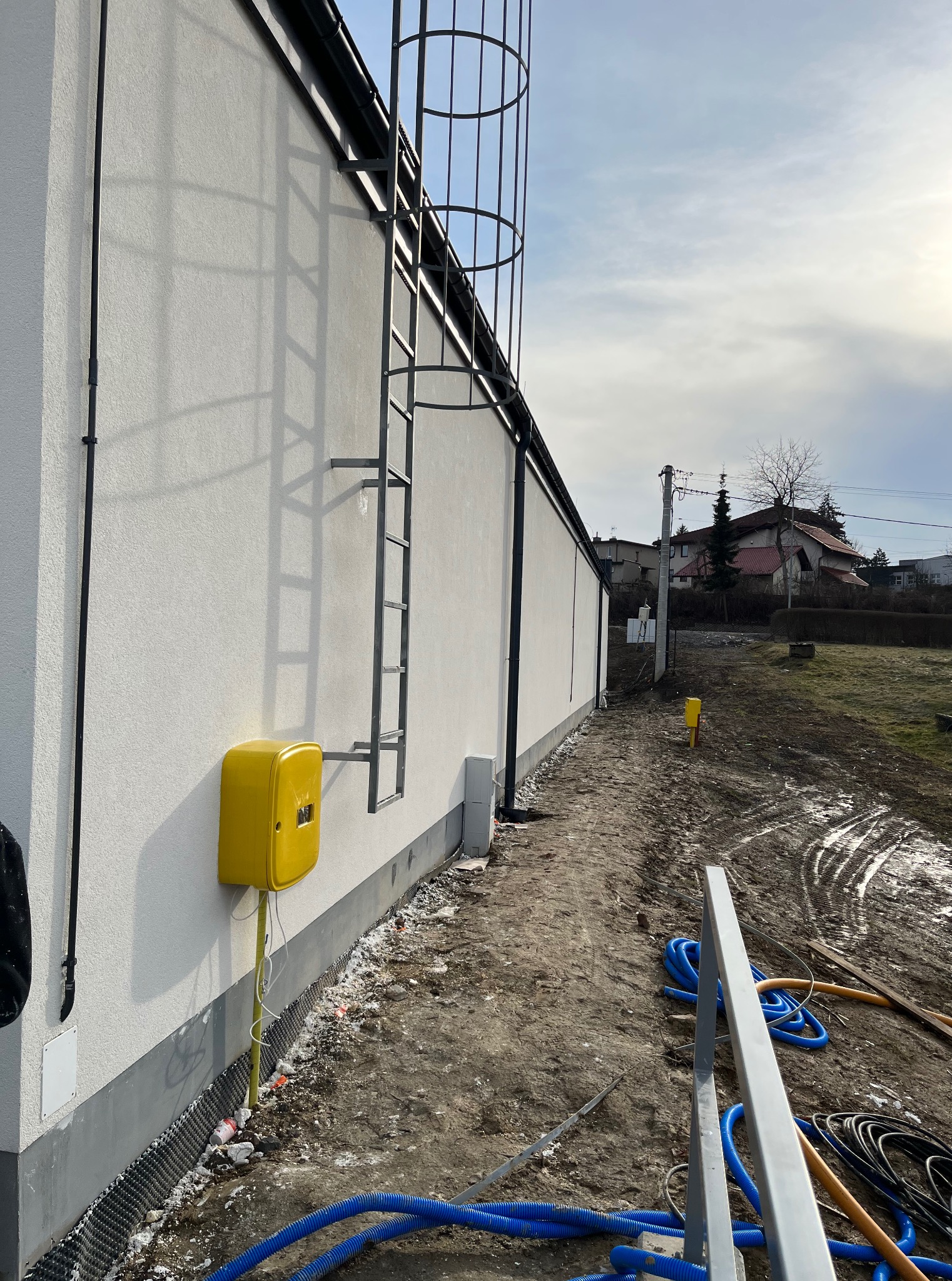 Biała elewacja budynku z żółtą skrzynką gazową i drabinką w Żywcu. Widoczne elementy instalacji i teren wokół budynku. Teksturowana powierzchnia elewacji.