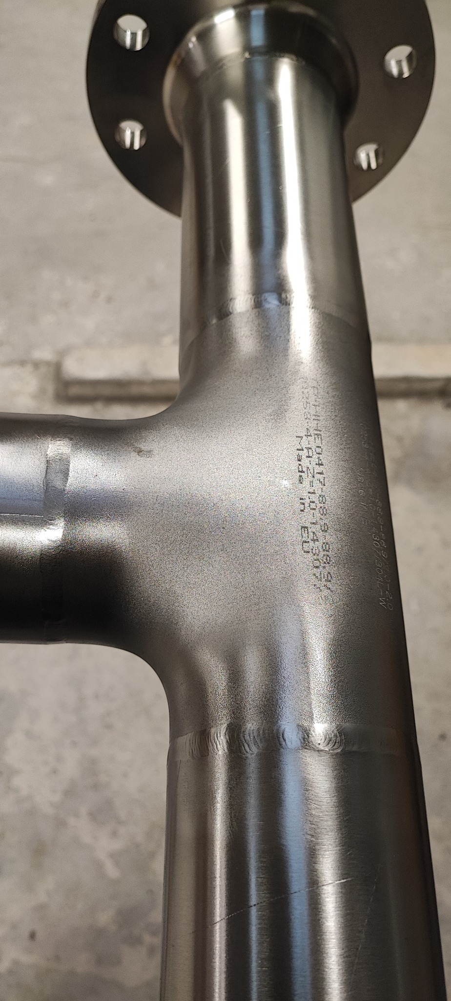 Spoina T ze stali nierdzewnej z kołnierzem, widoczne ślady po obróbce i napisy 'Made in EU'. Precyzyjne połączenia spawane.