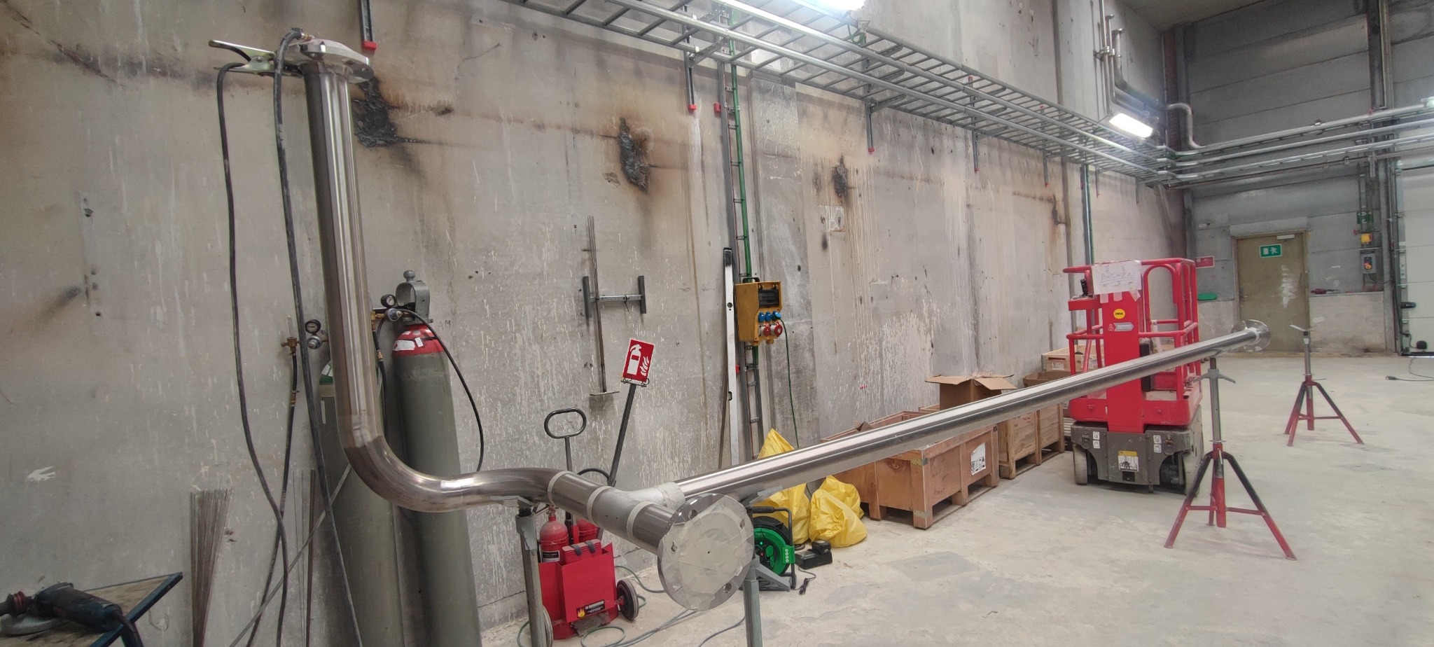 Spawanie rur ze stali nierdzewnej w hali przemysłowej, widoczny sprzęt spawalniczy, podnośnik i zabezpieczenia ppoż. Ślady spawania na ścianie.
