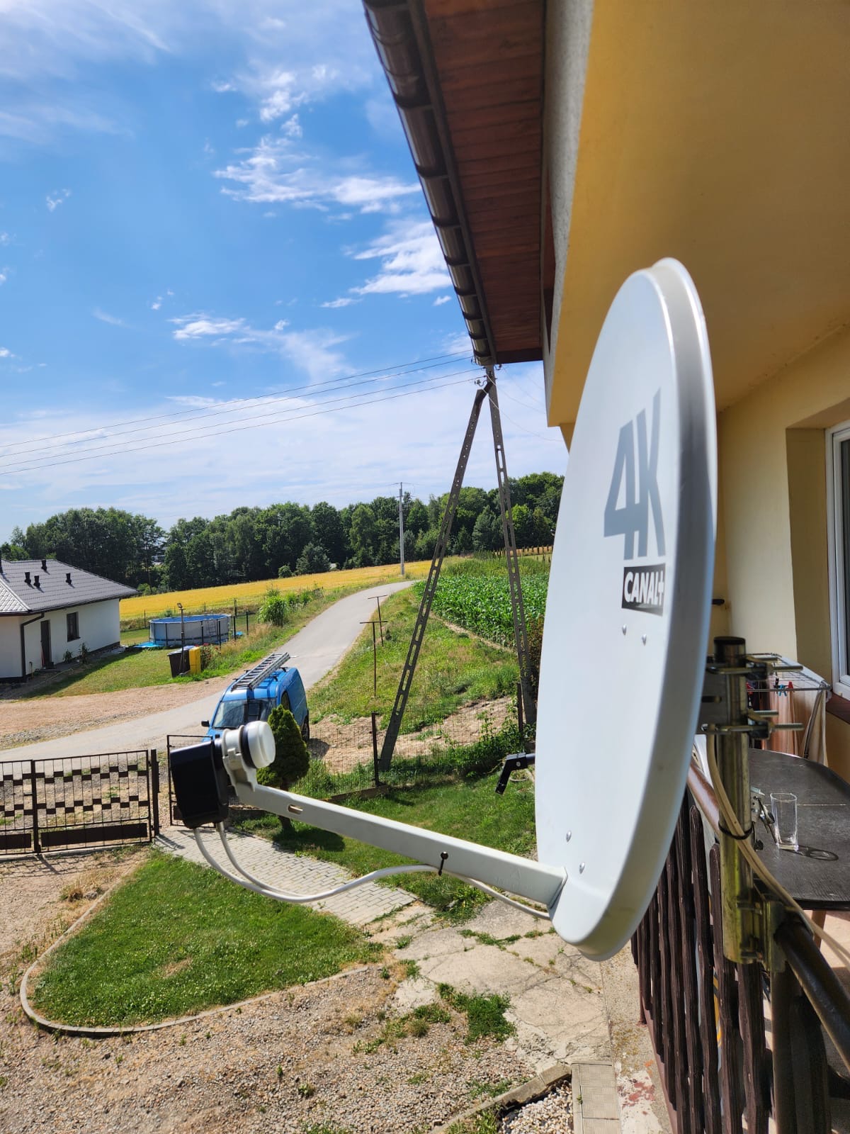 Biała antena satelitarna 4K Canal+ zamontowana na żółtej ścianie domu, na tle błękitnego nieba i zieleni. Na podwórku zaparkowany niebieski bus z drabiną.