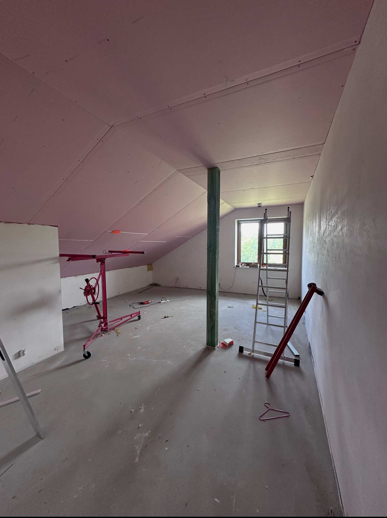 Poddasze w trakcie prac: różowa płyta karton-gips na skosach sufitu, metalowy podnośnik do płyt, drabina oparta o ścianę. Widoczne okno i słup konstrukcyjny.