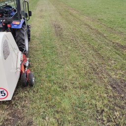 MarKop MARCIN KUCZMIERCZYK - Ciągnik z kosiarką rotacyjną na nierównym trawniku po koszeniu, widoczne ślady kół i skoszonej trawy. W tle drzewa i budynek.